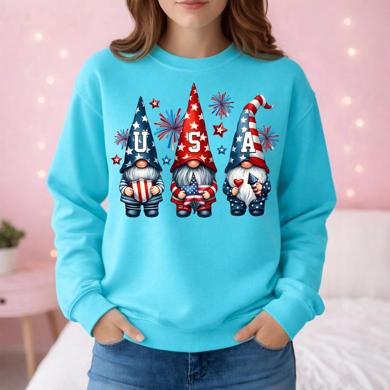 May include: A light blue sweatshirt featuring three gnomes with patriotic designs. Each gnome wears a tall hat with the letters "U", "S", and "A". The gnomes hold flags and fireworks are in the background. The sweatshirt is a solid light blue color.