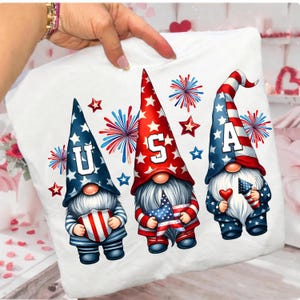 May include: White pillow with a patriotic design featuring three gnomes. Each gnome wears a tall hat with stars and stripes, holding a flag or heart. The word "USA" is spelled out across the hats. Fireworks and stars complete the design.