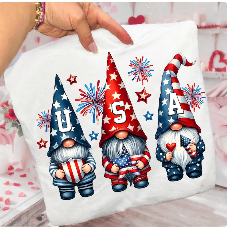 May include: White pillow with a patriotic design featuring three gnomes. Each gnome wears a tall hat with stars and stripes, holding a flag or heart. The word "USA" is spelled out across the hats. Fireworks and stars complete the design.