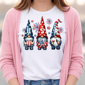 May include: White t-shirt featuring three patriotic gnomes with the letters "USA". The gnomes wear red, white, and blue hats and hold American flags and hearts. Fireworks and stars surround the gnomes, creating a festive design.