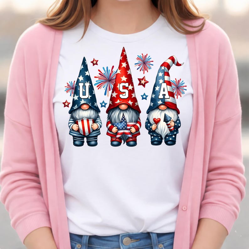 May include: White t-shirt featuring three patriotic gnomes with the letters "USA". The gnomes wear red, white, and blue hats and hold American flags and hearts. Fireworks and stars surround the gnomes, creating a festive design.