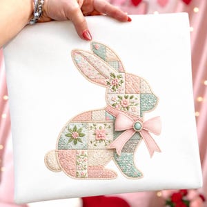 Może przedstawiać: Biała kwadratowa poduszka z patchworkowym wzorem kr&oacute;lika. Kr&oacute;lik składa się z pastelowych r&oacute;żowych, niebieskich i kremowych kwadrat&oacute;w z haftami kwiatowymi. R&oacute;żowa kokarda z turkusowym środkiem zdobi szyję kr&oacute;lika, tworząc motyw wielkanocny.