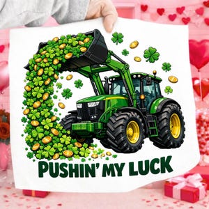 Pode incluir: Ilustração de um trator verde com uma pá cheia de trevos e moedas de ouro. O trator tem pneus pretos com jantes amarelas. O texto "PUSHIN' MY LUCK" está na parte inferior. A imagem está em um fundo branco.