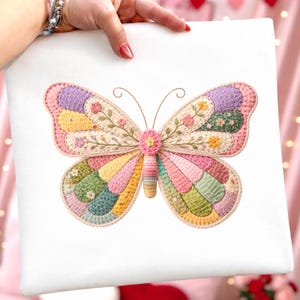 Może przedstawiać: Kolorowy, szydełkowy wz&oacute;r motyla na białym tle. Motyl ma patchwork z pastelowych kolor&oacute;w, w tym r&oacute;żowego, ż&oacute;łtego, zielonego i fioletowego. Delikatne haftowane kwiaty zdobią skrzydła, a korpus ma teksturowany, cylindryczny kształt.