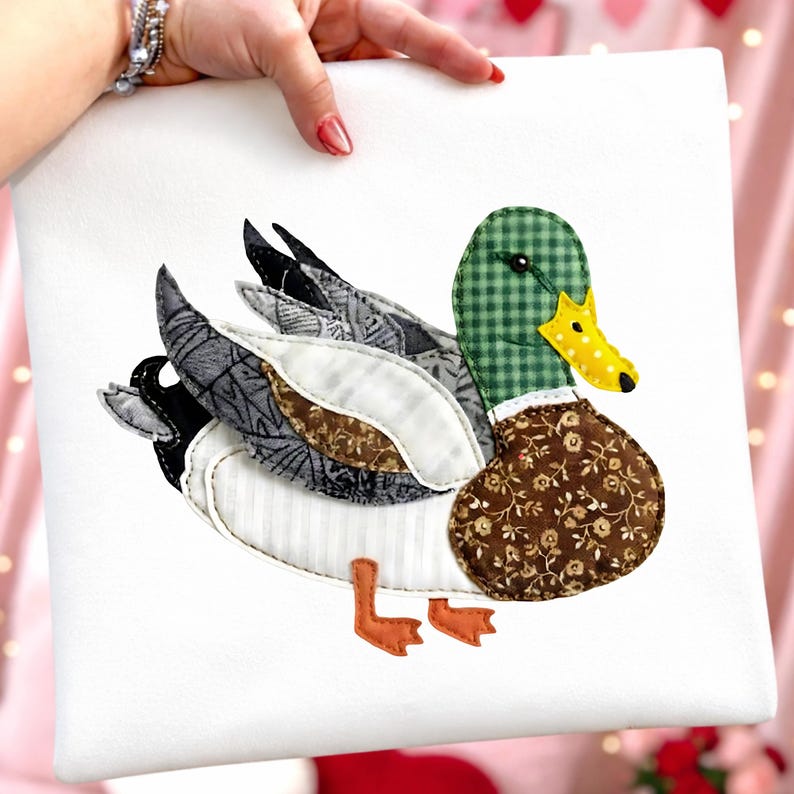 Może przedstawiać: Biała kwadratowa poduszka z aplikacją kaczki krzyż&oacute;wki. Kaczka ma zieloną głowę w kratkę, ż&oacute;łto-czarny kropkowany dzi&oacute;b i brązowy korpus we wzory kwiatowe. Skrzydła to mieszanka szarego, białego i czarnego materiału.