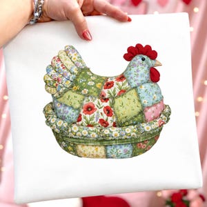 Może przedstawiać: Kolorowa, patchworkowa kura w gnieździe. Kura ma czerwony grzebień i dzi&oacute;b, a jej ciało składa się z r&oacute;żnych łatek z tkaniny kwiatowej. Gniazdo jest zielone ze wzorem stokrotek. Dzieło sztuki jest trzymane przez osobę.