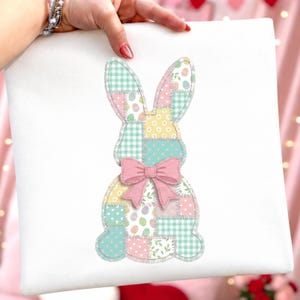 Może przedstawiać: Biała kwadratowa poduszka z patchworkowym wzorem kr&oacute;lika. Kr&oacute;lik składa się z r&oacute;żnych kwadrat&oacute;w w pastelowych kolorach, w tym kratki, groch&oacute;w i wzor&oacute;w kwiatowych. R&oacute;żowa kokarda jest umieszczona na środku klatki piersiowej kr&oacute;lika, dodając dekoracyjnego akcentu.