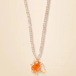 May include: A long necklace featuring a strand of small, translucent, pale pink beads. The necklace terminates in a vibrant, bright orange thread tassel that hangs from the center, creating a distinct contrast with the light-colored beaded chain.