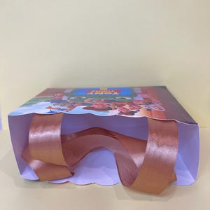 May include: A rectangular gift bag featuring a colorful graphic print of animated characters and the text TONY TURNS. The bag has a white interior and wide, metallic copper-colored satin ribbon handles attached to the top edges for carrying.