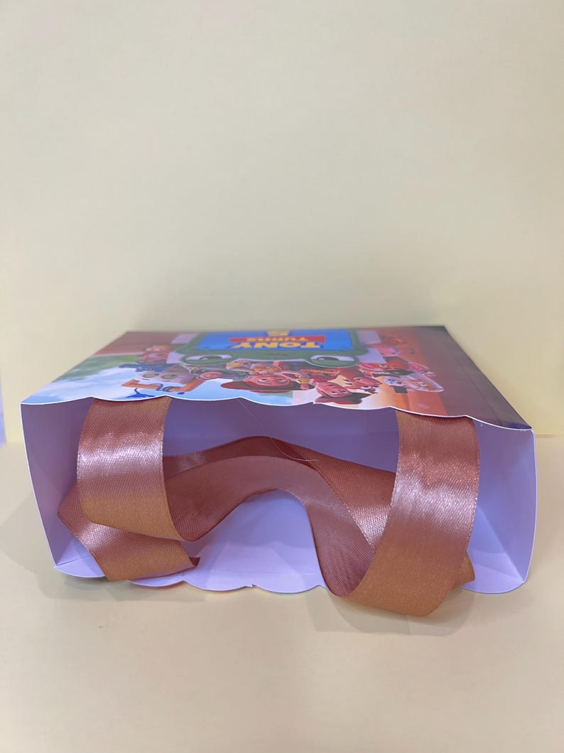 May include: A rectangular gift bag featuring a colorful graphic print of animated characters and the text TONY TURNS. The bag has a white interior and wide, metallic copper-colored satin ribbon handles attached to the top edges for carrying.