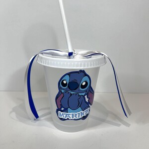 K&ouml;nnte beinhalten: Ein durchsichtiger Plastikbecher mit wei&szlig;em Deckel und Strohhalm. Der Becher zeigt eine blaue Cartoon-Figur mit dem Namen "Marina" darunter. Blaue und wei&szlig;e B&auml;nder sind um den Deckel gebunden.