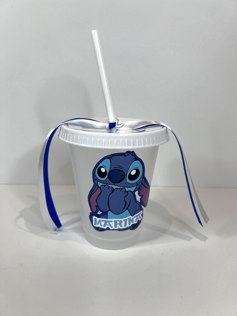 K&ouml;nnte beinhalten: Ein durchsichtiger Plastikbecher mit wei&szlig;em Deckel und Strohhalm. Der Becher zeigt eine blaue Cartoon-Figur mit dem Namen "Marina" darunter. Blaue und wei&szlig;e B&auml;nder sind um den Deckel gebunden.