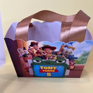 May include: A rectangular paper gift bag with scalloped top edges and brown satin ribbon handles. The front features a colorful Toy Story character graphic and a green frog-shaped frame with the text Tony Turns 5 in yellow and red lettering.