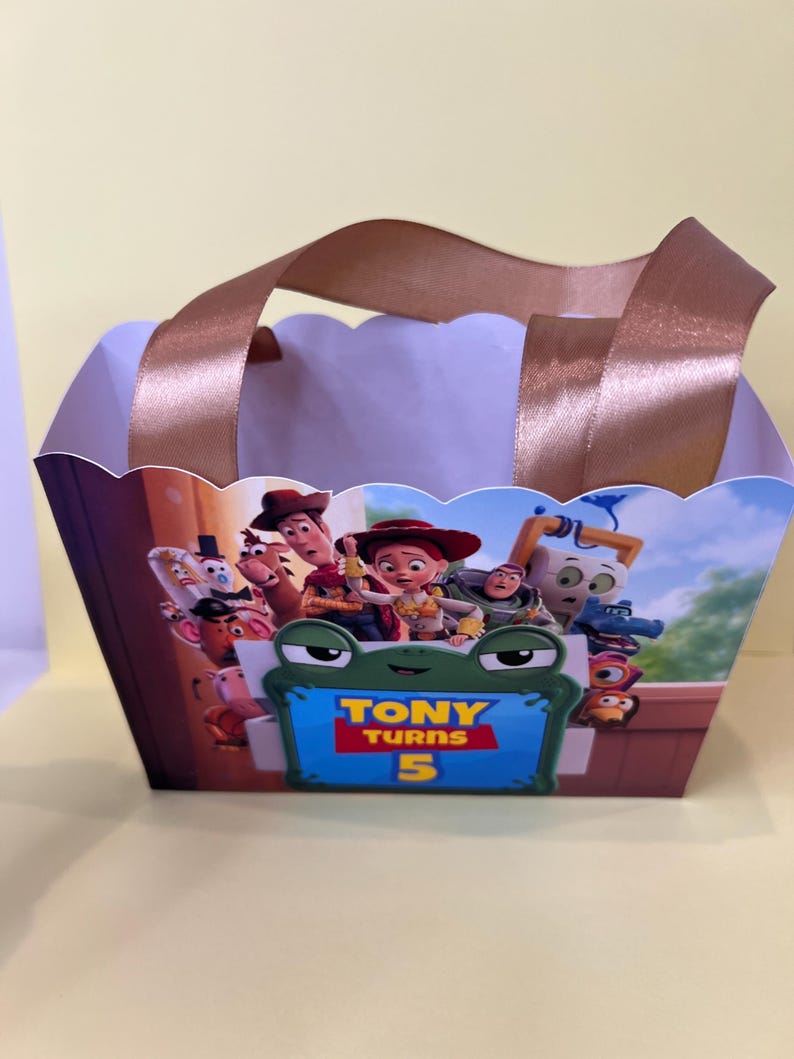May include: A rectangular paper gift bag with scalloped top edges and brown satin ribbon handles. The front features a colorful Toy Story character graphic and a green frog-shaped frame with the text Tony Turns 5 in yellow and red lettering.