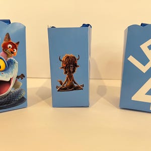 Pode incluir: Tr&ecirc;s caixas de presente retangulares azul-claras com bordas superiores recortadas. A caixa da esquerda apresenta uma cobra azul e uma raposa. A caixa central exibe uma criatura peluda em posi&ccedil;&atilde;o de medita&ccedil;&atilde;o. A caixa da direita mostra o texto branco LEG 4 TUR.