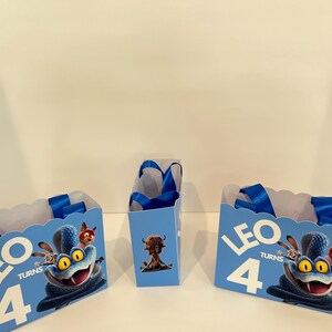 Pode incluir: Tr&ecirc;s sacos de presente azul-claros com bordas superiores recortadas e al&ccedil;as de fita azul. Os sacos apresentam um gr&aacute;fico de uma serpente azul com olhos amarelos, o texto LEO TURNS 4 e uma pequena criatura castanha na parte frontal.