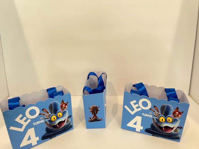 Pode incluir: Tr&ecirc;s sacos de presente azul-claros com bordas superiores recortadas e al&ccedil;as de fita azul. Os sacos apresentam um gr&aacute;fico de uma serpente azul com olhos amarelos, o texto LEO TURNS 4 e uma pequena criatura castanha na parte frontal.