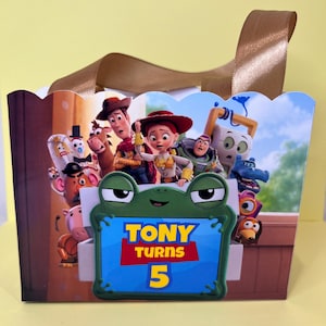 May include: A rectangular gift bag with a scalloped top edge and brown ribbon handles. The front features a colorful graphic of Toy Story characters and a central green frog-shaped frame containing the text TONY TURNS 5 in yellow and red.