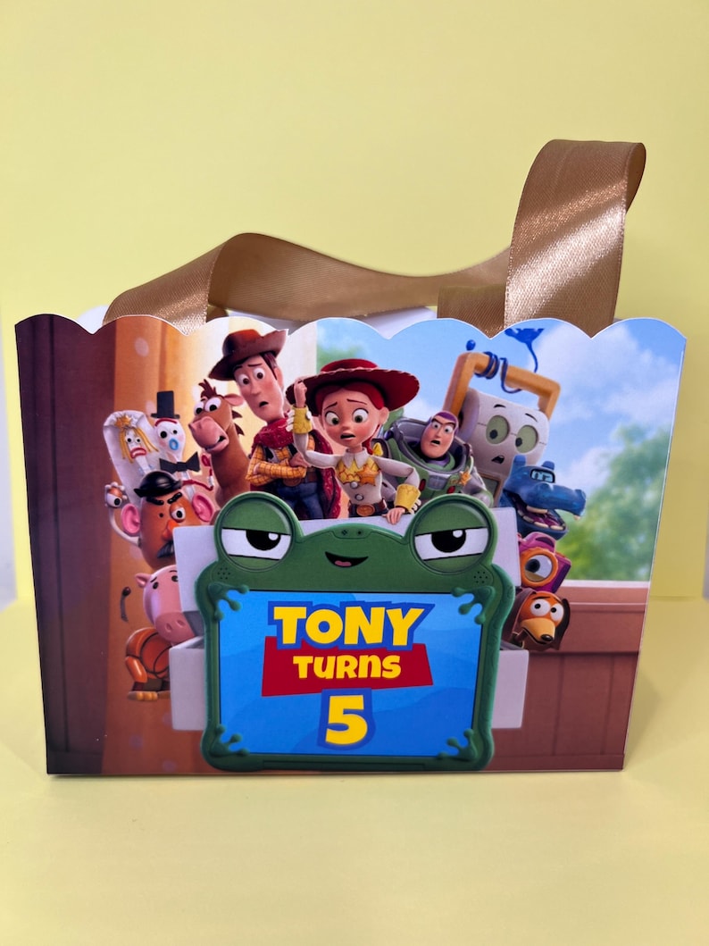 May include: A rectangular gift bag with a scalloped top edge and brown ribbon handles. The front features a colorful graphic of Toy Story characters and a central green frog-shaped frame containing the text TONY TURNS 5 in yellow and red.