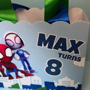 Puede incluir: Una bolsa de fiesta azul claro con ilustraciones de Spider-Man y Spider-Gwen. Tiene asas de cinta verde y azul y el texto "MAX TURNS 8". La parte inferior de la bolsa presenta un dise&ntilde;o de paisaje urbano.