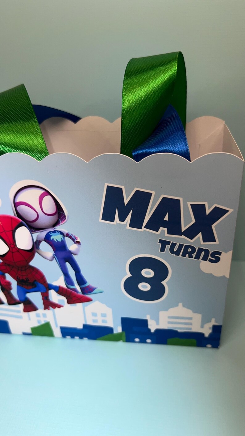 Puede incluir: Una bolsa de fiesta azul claro con ilustraciones de Spider-Man y Spider-Gwen. Tiene asas de cinta verde y azul y el texto "MAX TURNS 8". La parte inferior de la bolsa presenta un dise&ntilde;o de paisaje urbano.