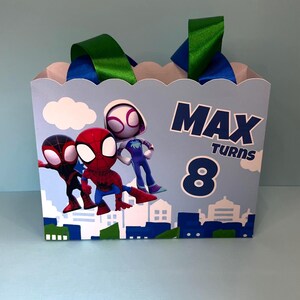 Puede incluir: Una caja de regalo azul claro con personajes de dibujos animados de Spider-Man. La caja tiene un borde festoneado, asas de cinta verde y azul, y el texto "MAX TURNS 8". Un dise&ntilde;o de paisaje urbano est&aacute; en la parte inferior.