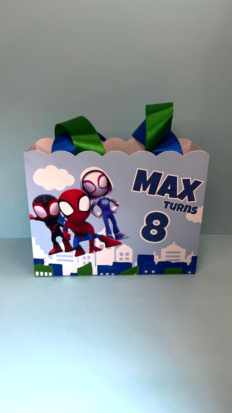 Puede incluir: Una caja de regalo azul claro con personajes de dibujos animados de Spider-Man. La caja tiene un borde festoneado, asas de cinta verde y azul, y el texto "MAX TURNS 8". Un dise&ntilde;o de paisaje urbano est&aacute; en la parte inferior.