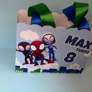 Puede incluir: Una caja de regalo azul claro con personajes de Spider-Man. La caja tiene asas de cinta verde y azul y el texto "MAX TURNS 8". La caja tiene un dise&ntilde;o de paisaje urbano.
