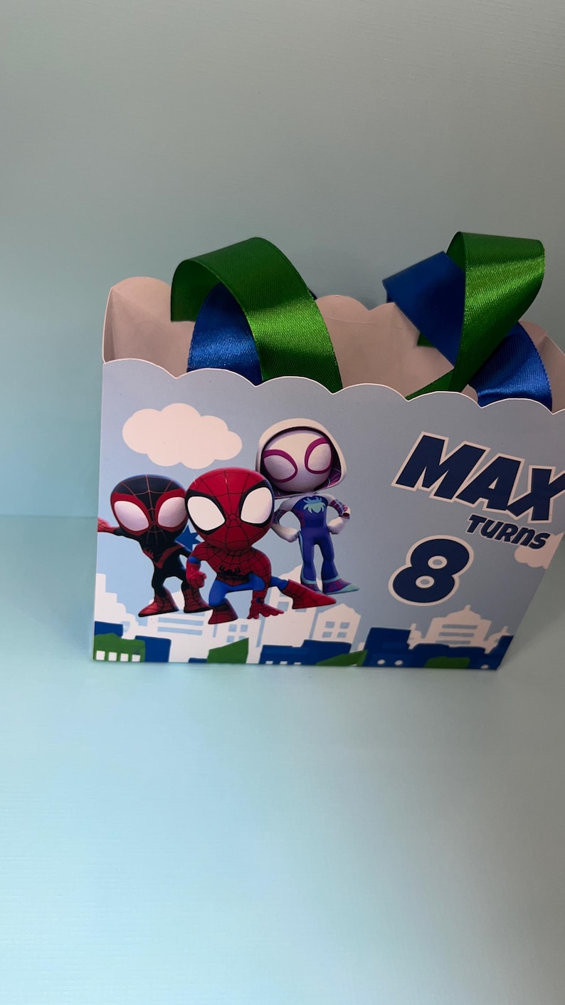 Puede incluir: Una caja de regalo azul claro con personajes de Spider-Man. La caja tiene asas de cinta verde y azul y el texto "MAX TURNS 8". La caja tiene un dise&ntilde;o de paisaje urbano.