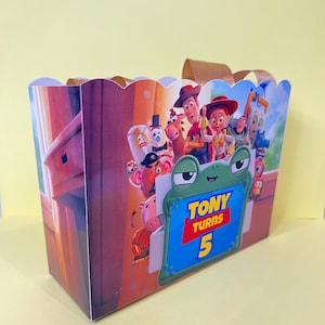 May include: A rectangular party favor box with a scalloped top edge and brown ribbon handles. The exterior features a colorful graphic of Toy Story characters and a green frog-shaped frame with the text Tony Turns 5 printed in yellow and red.