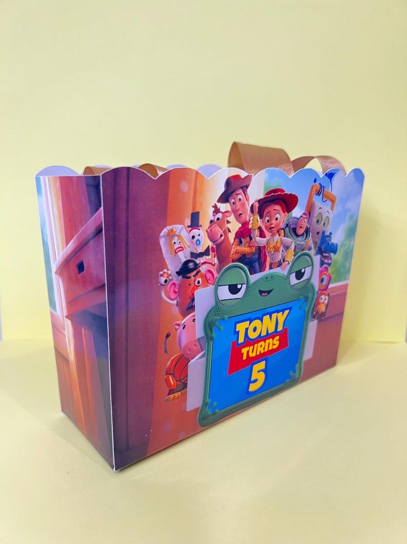May include: A rectangular party favor box with a scalloped top edge and brown ribbon handles. The exterior features a colorful graphic of Toy Story characters and a green frog-shaped frame with the text Tony Turns 5 printed in yellow and red.