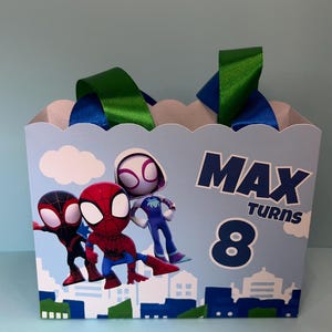Puede incluir: Una caja de regalo azul claro con una parte superior festoneada, con personajes de Spider-Man y el texto "MAX TURNS 8". La caja tiene asas de cinta verde y azul, ideal para una celebraci&oacute;n de cumplea&ntilde;os.
