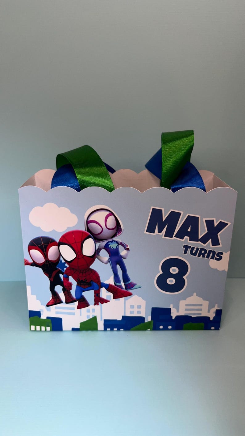 Puede incluir: Una caja de regalo azul claro con una parte superior festoneada, con personajes de Spider-Man y el texto "MAX TURNS 8". La caja tiene asas de cinta verde y azul, ideal para una celebraci&oacute;n de cumplea&ntilde;os.