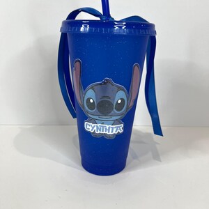 K&ouml;nnte beinhalten: Blauer Becher mit Deckel, Strohhalm und Schleife. Der Becher zeigt ein Cartoon-Bild von Stitch und den Namen "CYNTHIA" in Wei&szlig;. Der Becher hat eine blaue Grundfarbe mit einem gesprenkelten Design.