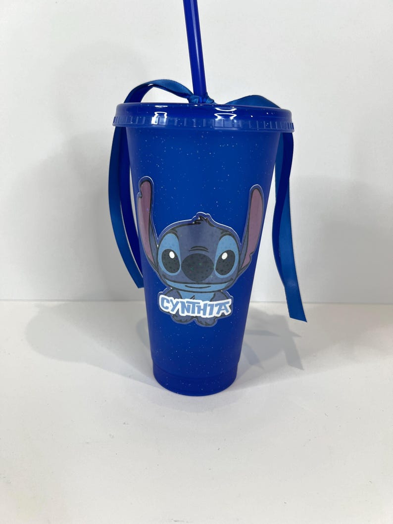K&ouml;nnte beinhalten: Blauer Becher mit Deckel, Strohhalm und Schleife. Der Becher zeigt ein Cartoon-Bild von Stitch und den Namen "CYNTHIA" in Wei&szlig;. Der Becher hat eine blaue Grundfarbe mit einem gesprenkelten Design.
