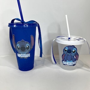 K&ouml;nnte beinhalten: Zwei Plastikbecher mit Deckeln und Strohhalmen. Einer ist blau mit einem Stitch-Motiv und dem Namen "CYNTHIA" in Wei&szlig;. Der andere ist transparent mit einem Stitch-Motiv und dem Namen "MARINA" in Wei&szlig;. Beide haben blaue B&auml;nder an den Deckeln.