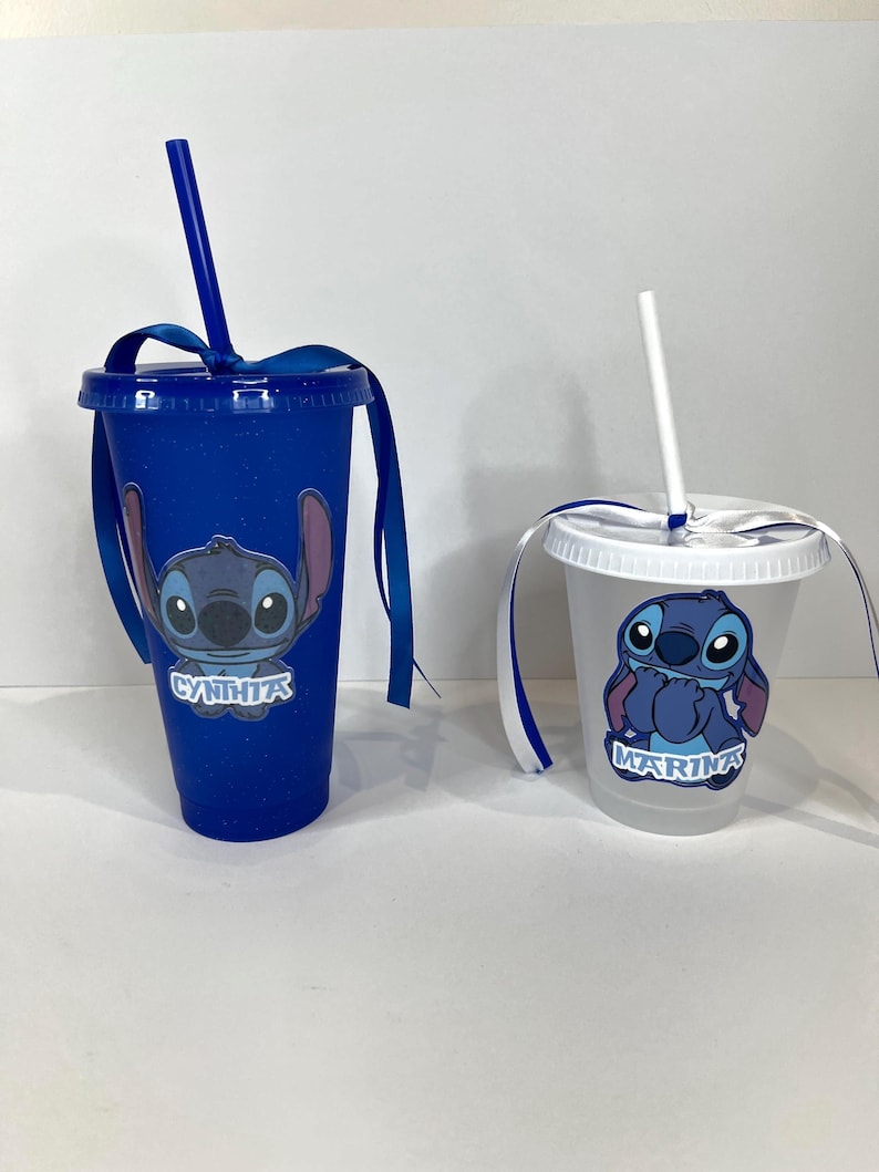 K&ouml;nnte beinhalten: Zwei Plastikbecher mit Deckeln und Strohhalmen. Einer ist blau mit einem Stitch-Motiv und dem Namen "CYNTHIA" in Wei&szlig;. Der andere ist transparent mit einem Stitch-Motiv und dem Namen "MARINA" in Wei&szlig;. Beide haben blaue B&auml;nder an den Deckeln.
