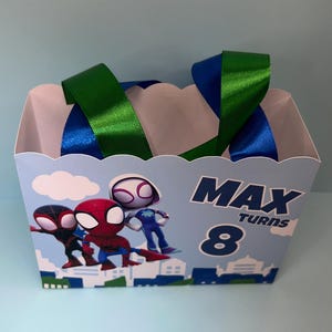 Puede incluir: Una caja de recuerdos de fiesta azul claro con personajes de Spider-Man. La caja tiene asas de cinta verde y azul. El texto en la caja dice "MAX TURNS 8". La caja est&aacute; decorada con im&aacute;genes de dibujos animados de Spider-Man.