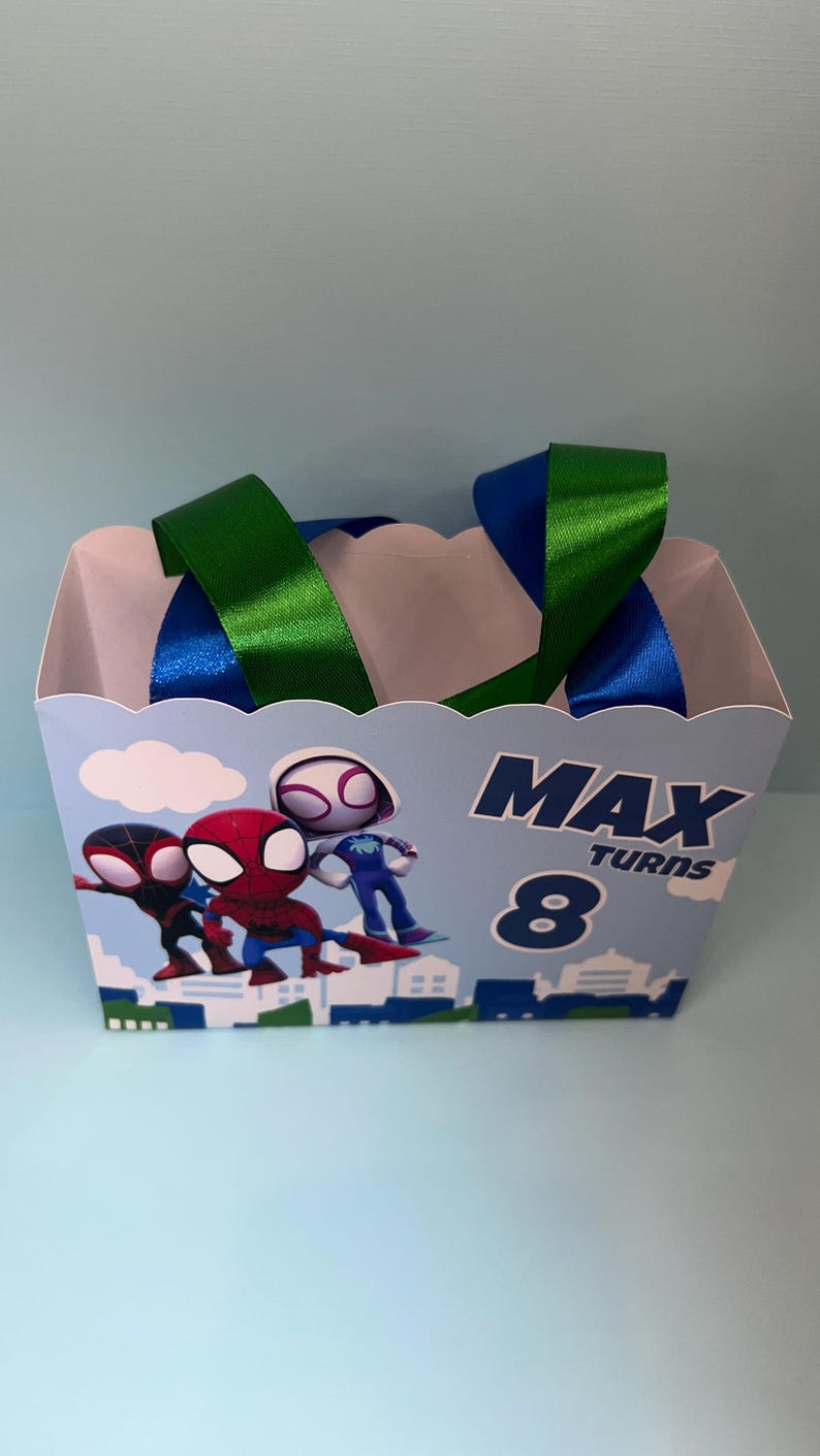 Puede incluir: Una caja de recuerdos de fiesta azul claro con personajes de Spider-Man. La caja tiene asas de cinta verde y azul. El texto en la caja dice "MAX TURNS 8". La caja est&aacute; decorada con im&aacute;genes de dibujos animados de Spider-Man.