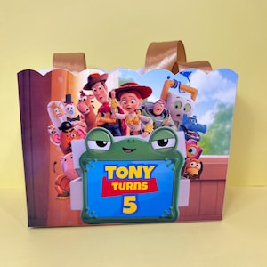May include: A rectangular gift bag with gold ribbon handles featuring a colorful Toy Story character print. A central green frog-shaped frame displays the text TONY TURNS 5 in yellow and red against a blue background, surrounded by various movie characters.