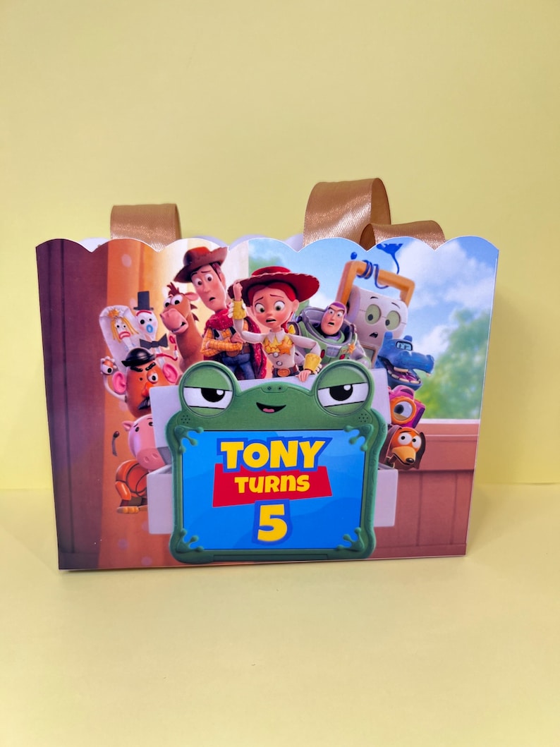 May include: A rectangular gift bag with gold ribbon handles featuring a colorful Toy Story character print. A central green frog-shaped frame displays the text TONY TURNS 5 in yellow and red against a blue background, surrounded by various movie characters.
