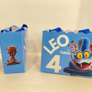 Pode incluir: Sacos de presente azul-claros com bordas superiores recortadas e al&ccedil;as de fita azul. A frente apresenta uma grande cobra azul, um coelho e uma raposa com o texto branco LEO TURNS 4. O painel lateral exibe uma criatura peluda castanha sentada.