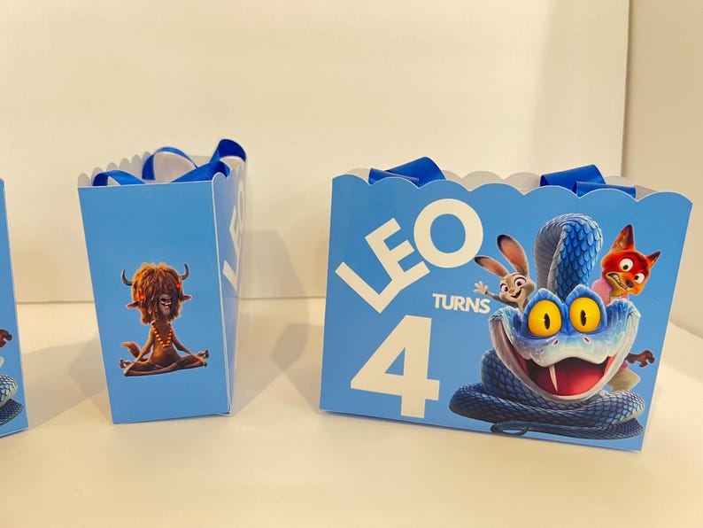 Pode incluir: Sacos de presente azul-claros com bordas superiores recortadas e al&ccedil;as de fita azul. A frente apresenta uma grande cobra azul, um coelho e uma raposa com o texto branco LEO TURNS 4. O painel lateral exibe uma criatura peluda castanha sentada.