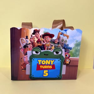 May include: A rectangular party favor bag featuring a colorful graphic of Toy Story characters and a central green frog-shaped frame with the text Tony Turns 5. The bag has a scalloped top edge and two brown ribbon handles attached to the back.