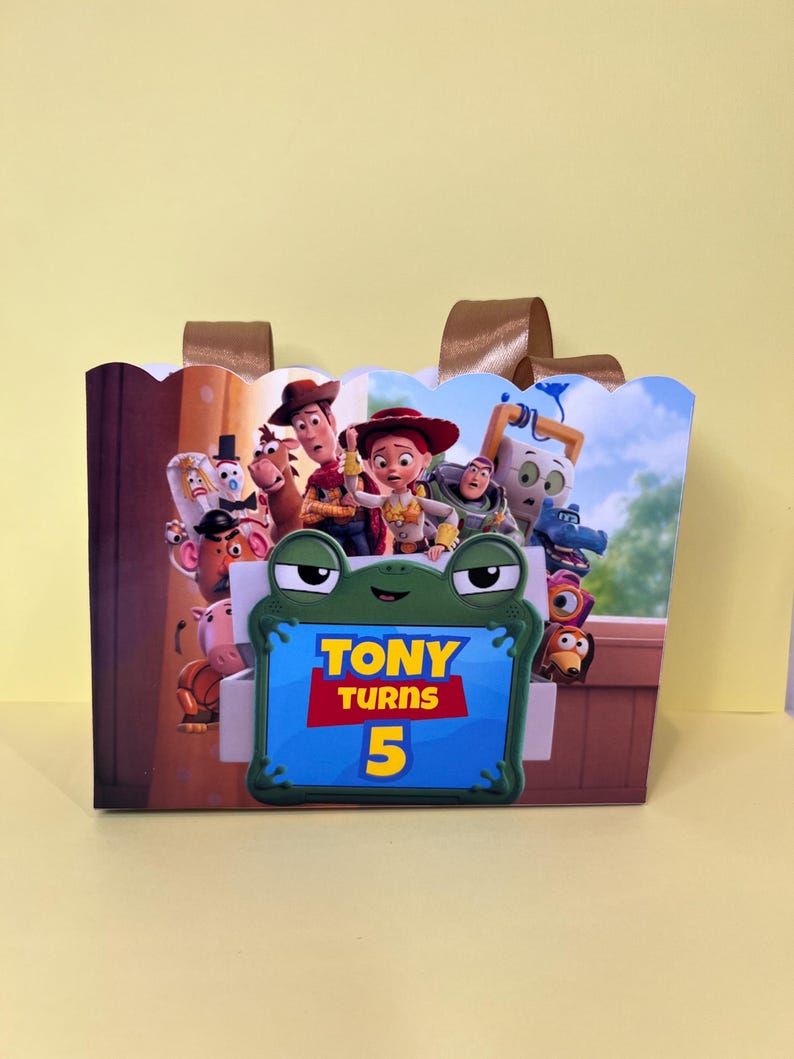 May include: A rectangular party favor bag featuring a colorful graphic of Toy Story characters and a central green frog-shaped frame with the text Tony Turns 5. The bag has a scalloped top edge and two brown ribbon handles attached to the back.