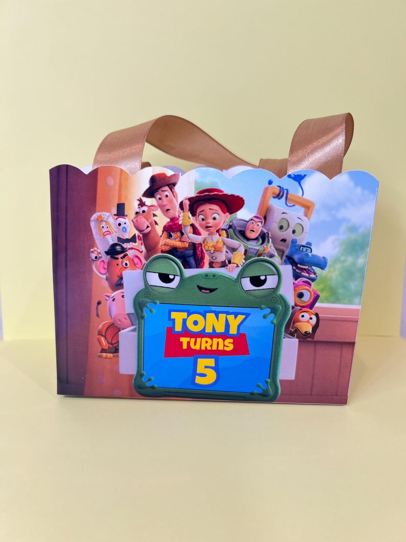May include: A rectangular gift bag with brown ribbon handles featuring a Toy Story character graphic and a central green frog-shaped sign with the text TONY TURNS 5 in yellow and red lettering against a blue background on the front panel.