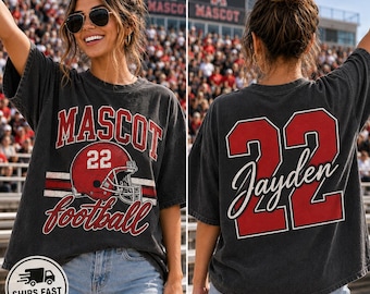 Puede incluir: Una camiseta gris carbón de manga corta con un gráfico de casco de fútbol americano rojo con el número 22 y el texto MASCOT football en el frente. La parte posterior muestra el número 22 grande en rojo con el nombre Jayden en letra blanca.