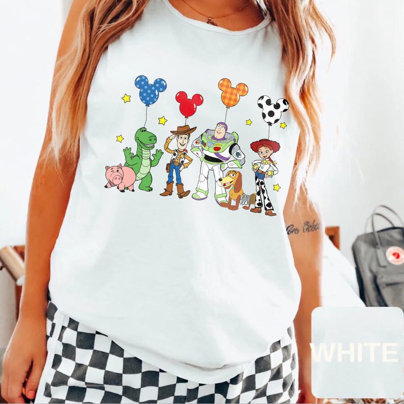 May include: White tank top featuring a colorful graphic of Toy Story characters holding Mickey Mouse-shaped balloons. The design includes Woody, Buzz Lightyear, Jessie, Rex, Hamm, and Slinky Dog, with yellow star accents.