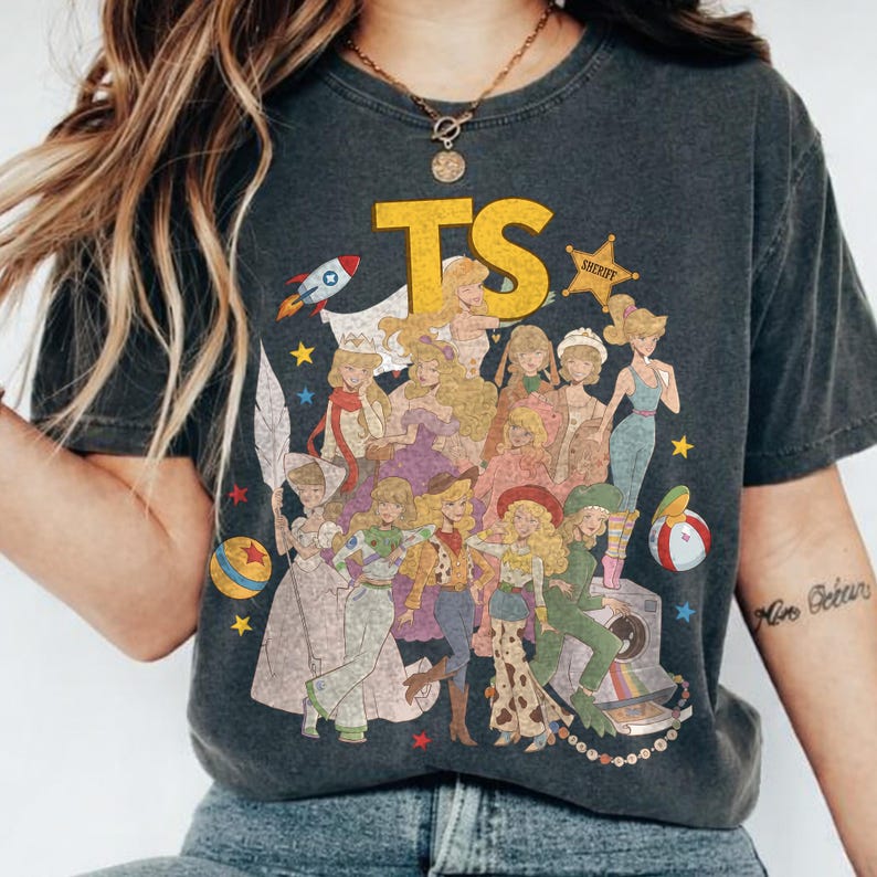 May include: A charcoal gray short-sleeve t-shirt featuring a large yellow TS graphic and a collage of illustrated characters dressed in various outfits, including a space suit, cowboy attire, and gowns, surrounded by stars, a rocket, and a beach ball.