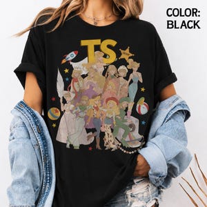 May include: A black t-shirt featuring a large graphic print of multiple animated characters in various outfits, including a space suit, cowboy attire, and formal gowns. The text TS is printed in large yellow letters at the top, with a sheriff star badge.